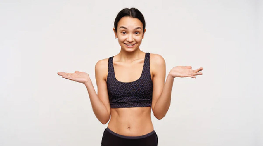 Young dark haired girl shrugging in a black sports bra with her palms open in front or a pale grey background