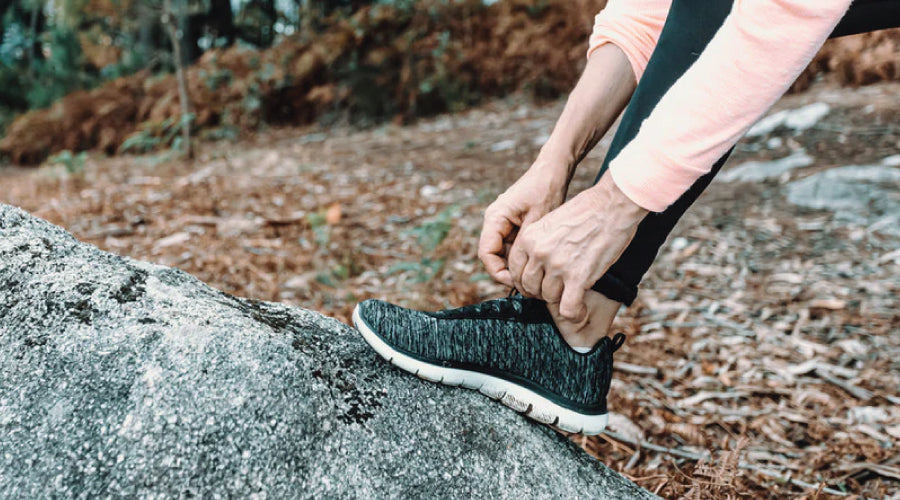 Woman outdoors tying her trainer shoe whilst leaning it again a rock in the woods