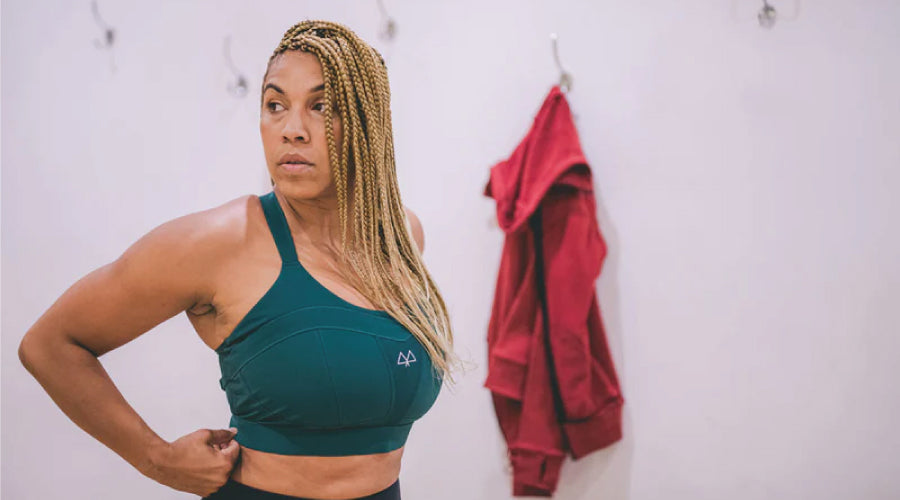 Strong woman in a changing room wearing a green Maaree sports bra with her red hoodie hanging on a peg