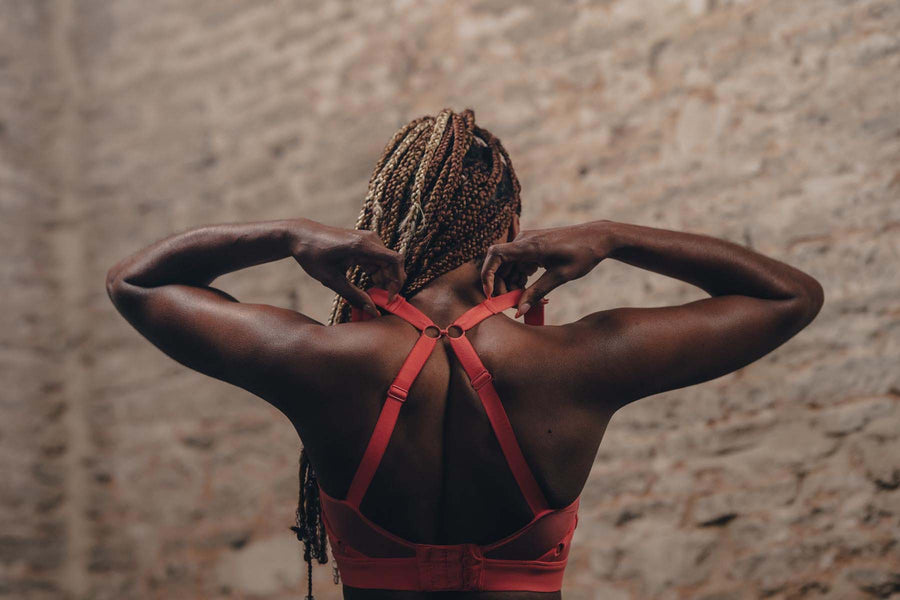 A woman stands in the gym with her back to the camera, she is adjusting the racerback on her coral sports bra.